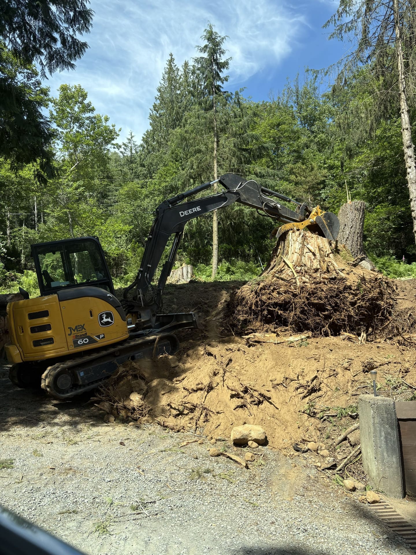 A JMEX John Deere 60P excavator lifting a cleared stump and root ball on a wooded Fraser Valley site