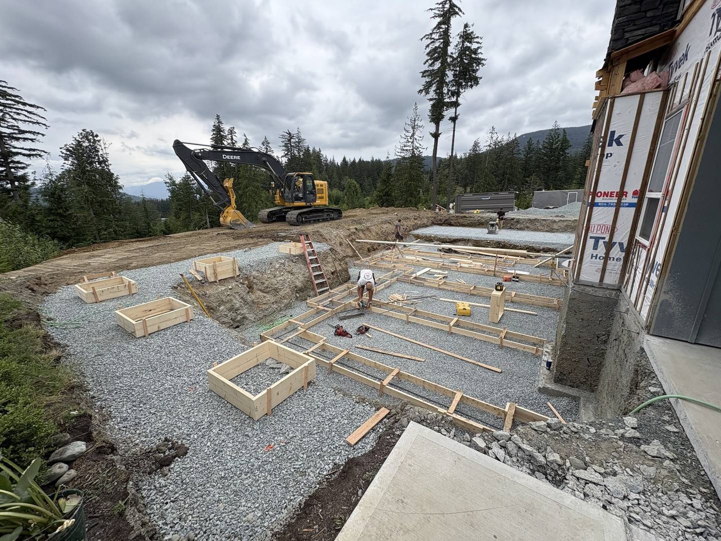 Formed concrete footings on a gravel pad beside a house under construction, with an excavator working on the bench above