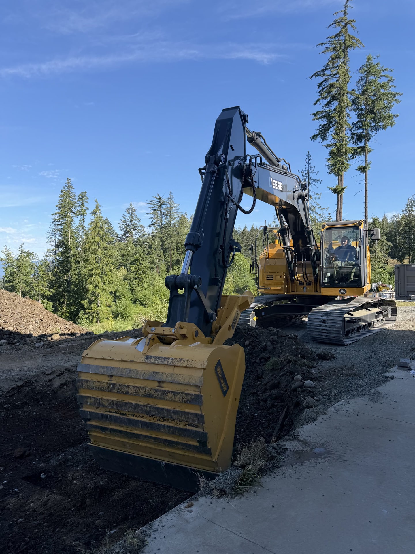 A tracked John Deere excavator with its bucket lowered to a bank of dark soil beside a new concrete edge, the operator in the cab, tall firs and open sky behind