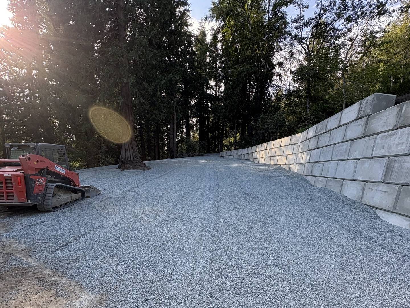 A concrete block retaining wall running along the edge of a newly built gravel driveway, with a compact track loader parked at the treeline