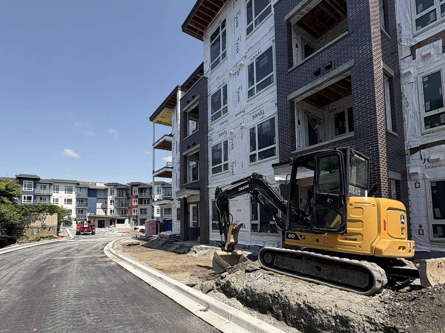 A finished asphalt access road curving past a multi-family building under construction, with a compact excavator working the shoulder