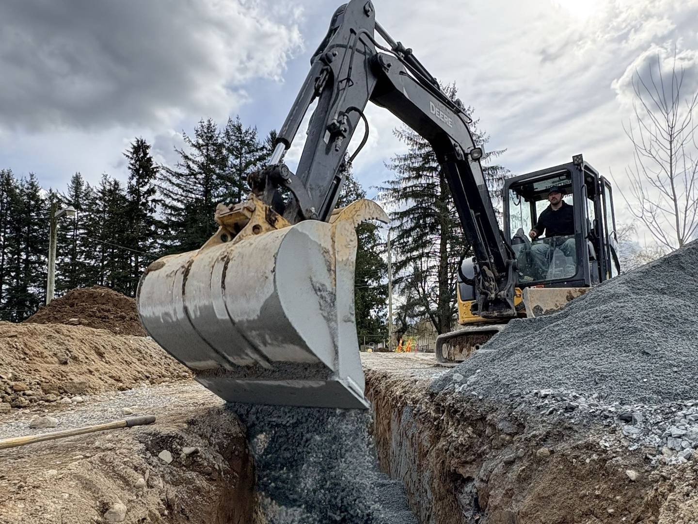An excavator bucket releasing bedding gravel into an open trench with service pipe already laid in the bottom