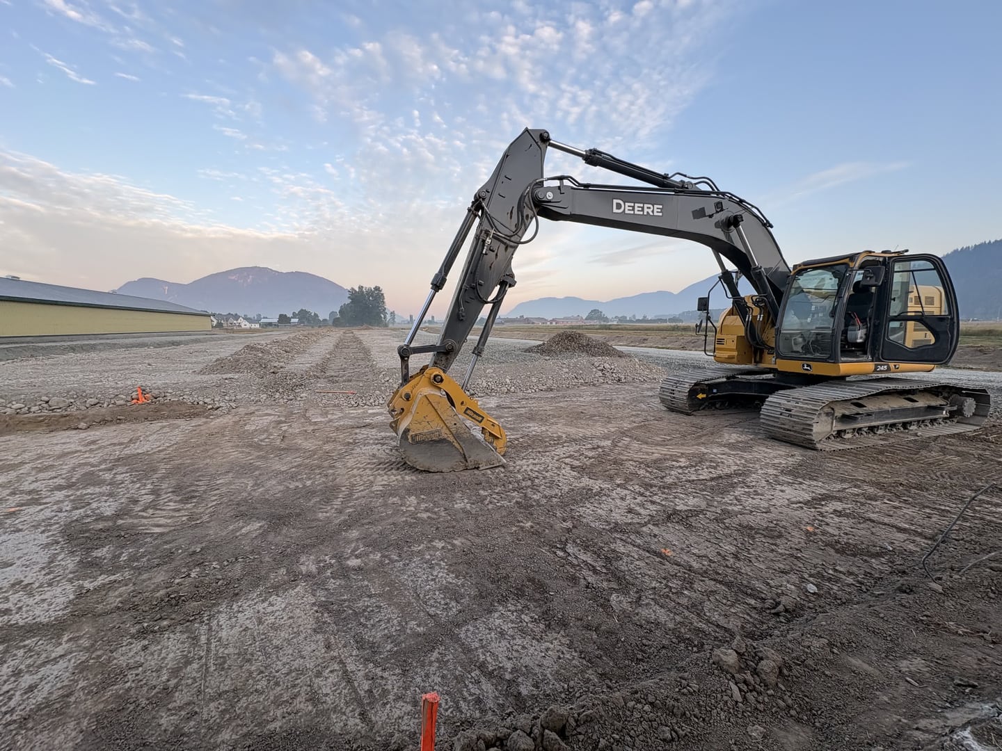 A JMEX excavator at golden hour on a freshly graded Fraser Valley site, spoil rows behind it and the mountains on the horizon