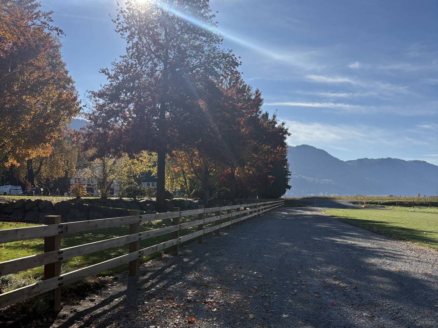 A gravel driveway running beside a post-and-rail fence, autumn sun through the trees and a mountain ridge behind