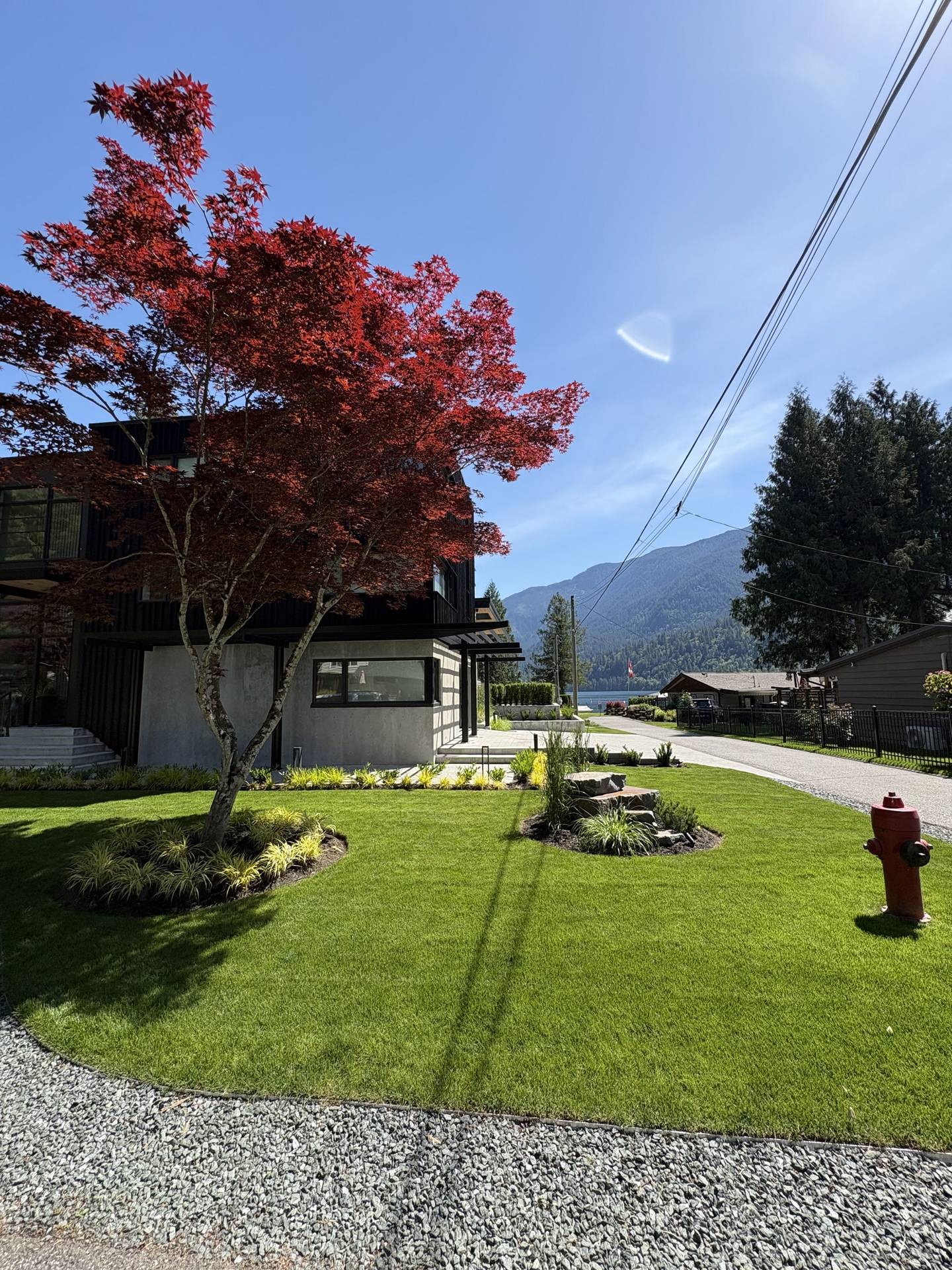 A finished lawn and planted beds around a modern house, with a red Japanese maple, a stone feature bed and the lake and mountains behind