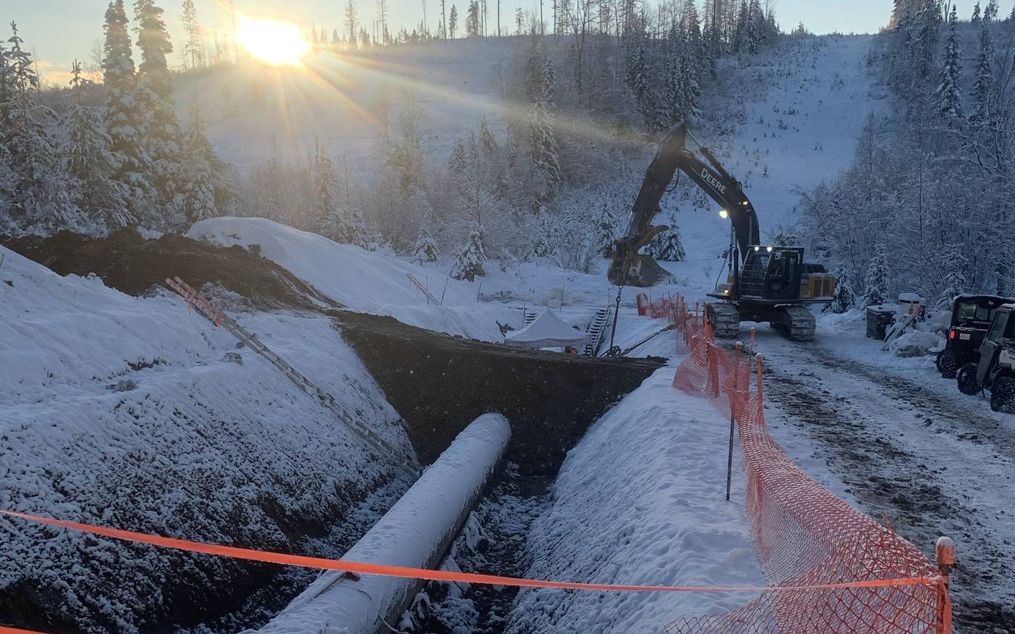 A snow-covered pipe along an open trench, an excavator working beyond it, orange safety fencing at the edge and a low winter sun over the trees