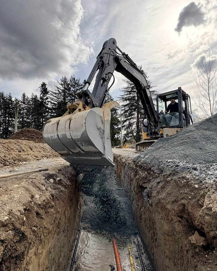 An excavator bucket over an open trench with two service pipes laid in the bottom of it, the operator at work in the cab
