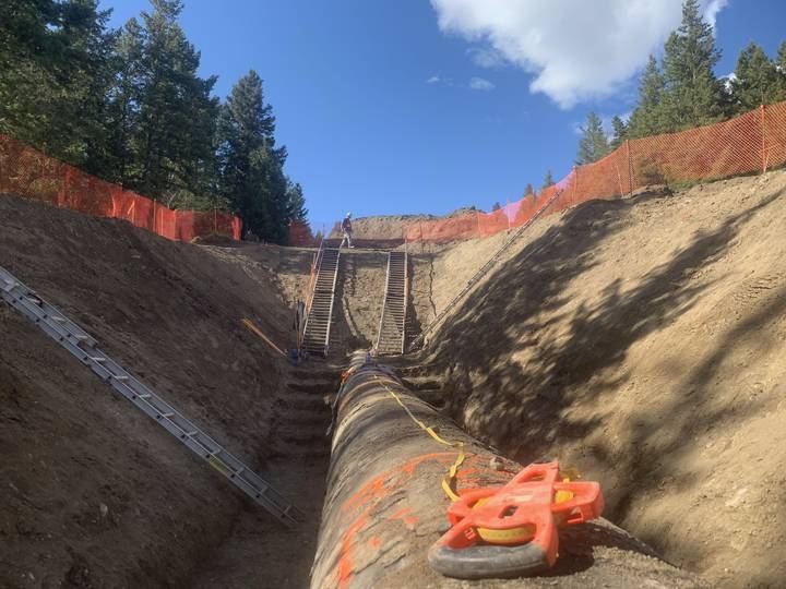 Looking along the top of a large pipe in the bottom of a deep trench, with two sets of stairs and ladders up the far wall and a worker standing at the top