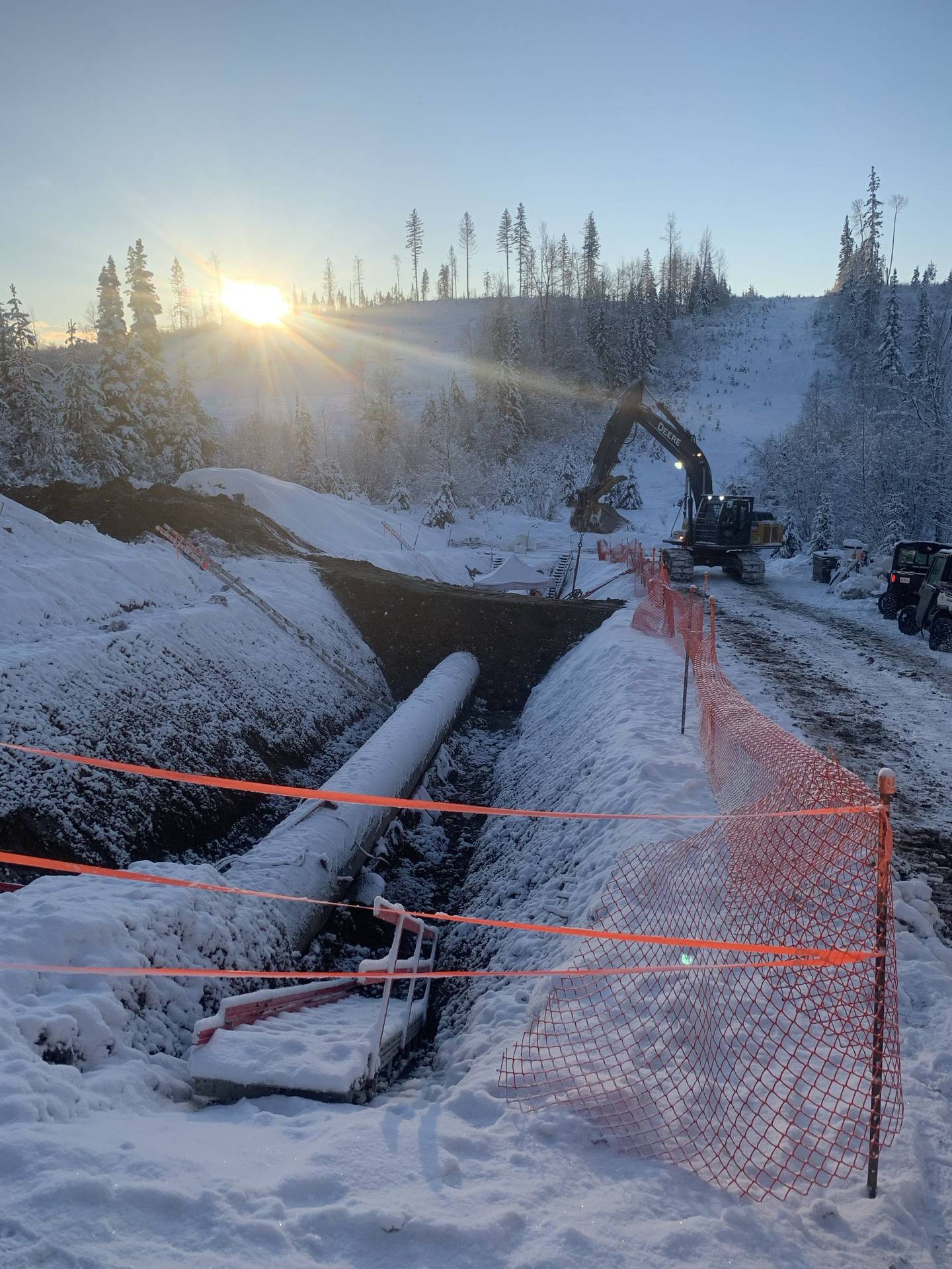 A snow-covered pipe along an open trench, an excavator working beyond it, orange safety fencing at the edge and a low winter sun over the trees