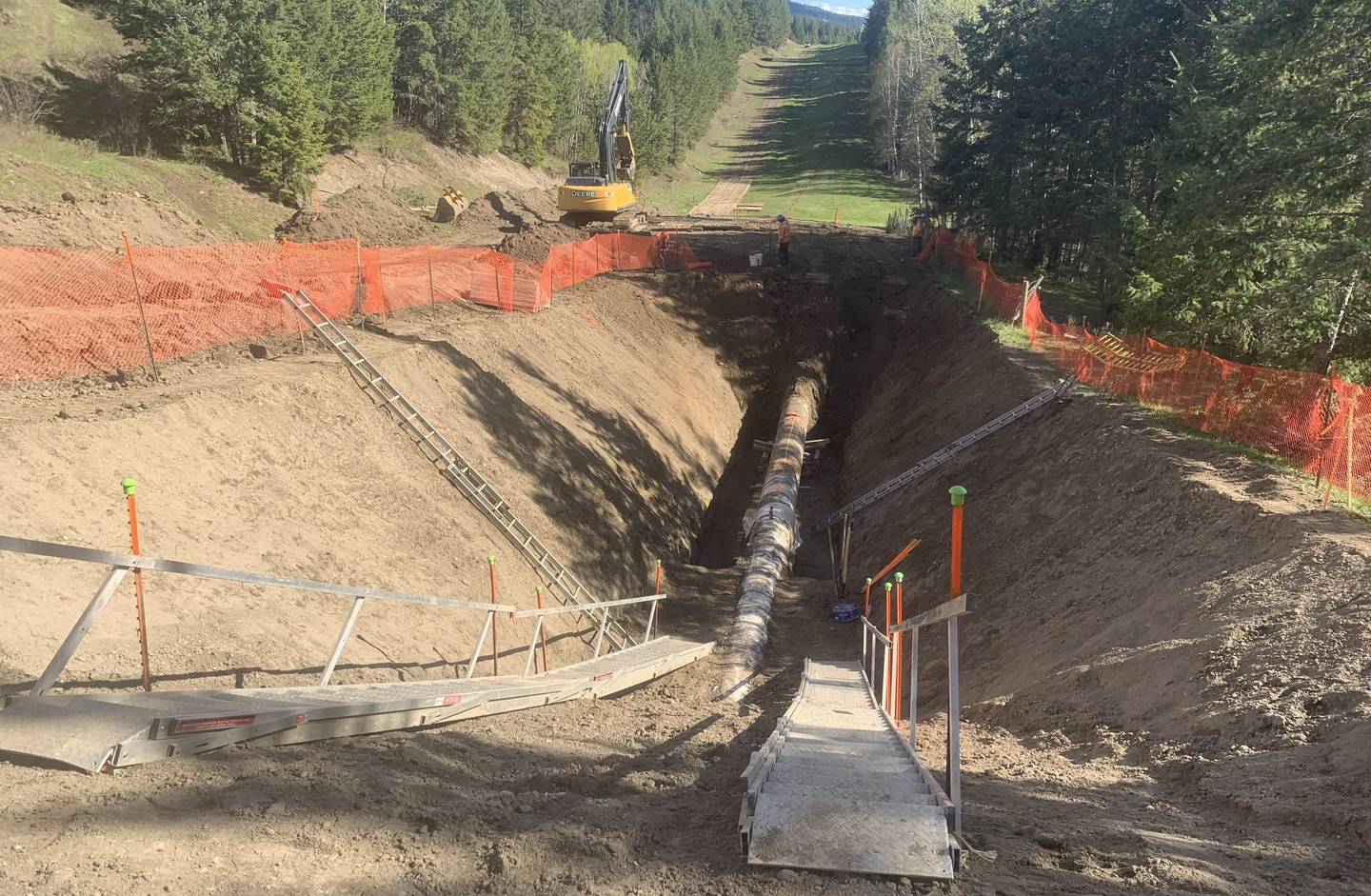 A deep trench with a large pipe in the bottom, stairs and a walkway leading down into it, ladders on the walls and an excavator at the far end